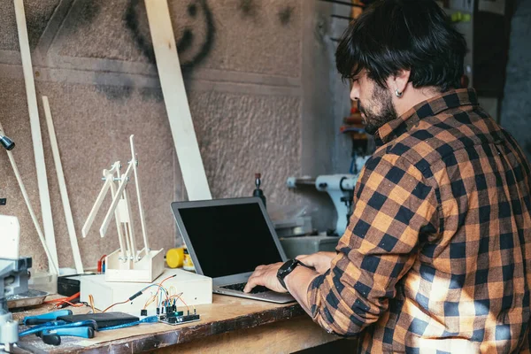 Mühendis atölyesinde dizüstü bilgisayar kullanıyor. Robot dükkanında çalışırken dizüstü bilgisayarda bir şey yazan ve robot kolu yapan ciddi bir adam..