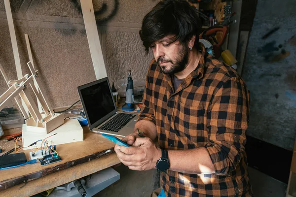 Mühendis atölyesinde cep telefonu kullanıyor. Ciddi bir adam akıllı telefonuna mesaj yazarken çalışma masasında tahta robot kol yapıyor..