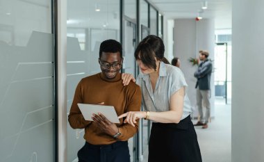 Mutlu İş Kadını ve İş Adamı İş yerinde Dijital Tablet Kullanıyor. İki gülümseyen meslektaşım, arka planda tanınmayan iki iş arkadaşıyla modern bir koridorda dururken bir tabletin iş raporunu inceliyor..