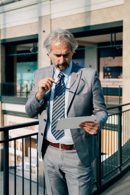 Ofis koridorunda dikilirken Dijital Tablet kullanan Ciddi İşadamı. Zarif gri takım elbiseli olgun iş adamı modern koridorda dururken tabletinde iş raporunu okuyor..