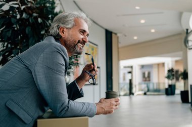 Kahve molası: Gülümseyen iş adamı iş yerinde bir fincan kahve içiyor. Modern ofis binasında bir masaya yaslanırken elinde çay ve gözlük tutan zarif gri takım elbiseli mutlu bir iş adamı..