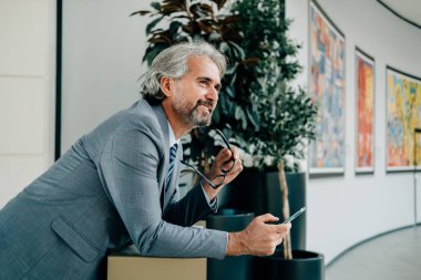 Gülümseyen Olgun İş Adamı iş yerinde cep telefonu kullanıyor. Modern ofis koridorunda bir masaya yaslanıp bir şeyler düşünürken zarif gri takım elbiseli mutlu bir iş adamı telefonunu ve gözlüklerini tutuyor..