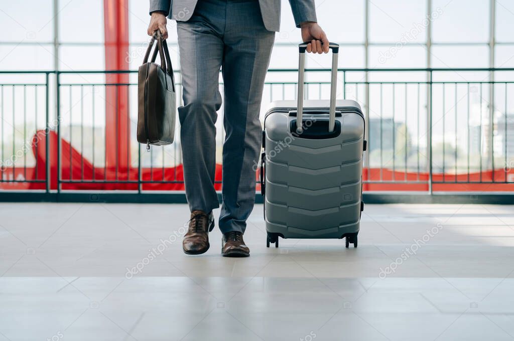 An Anonymous Business Man Walking With Luggage in the Airport. Close up photo of unrecognizable businessman in gray suit walking through airport terminal with briefcase and suitcase.