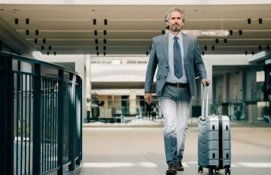 Havaalanında Bavulla Yürüyen Olgun İş Adamı 'nız kutlu olsun. Zarif gri takım elbiseli gülümseyen işadamı havaalanı terminalinde bavuluyla yürüyor..