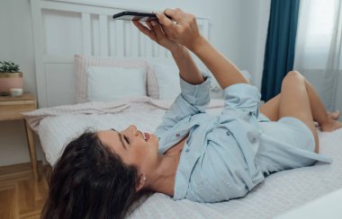 Gömlekli güzel bir kadın yatakta uzanıyor ve mesaj yazıyor ya da sabahları telefonundan bir şey izliyor.