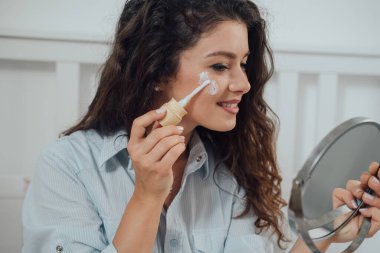 Gülümseyen kadının yatak odasında aynayı tutarken yüzüne krem sürüşünün yakın plan fotoğrafı.