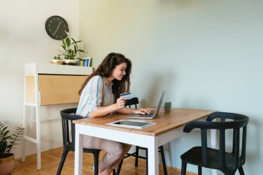 Gülümseyen İş kadını İnternetten Kredi kartını ve bilgisayarını kullanarak alışveriş yapıyor. Mutlu iş kadını, oturma odasında bir fincan kahve ve dijital tabletle masada otururken dizüstü bilgisayar ve kredi kartını kullanıyor..