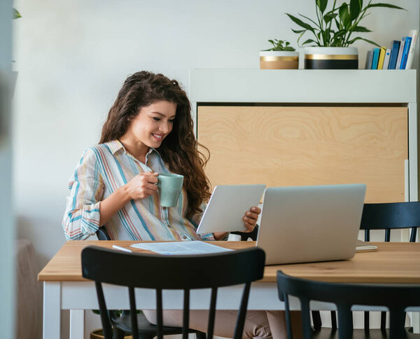 Счастливая деловая женщина, использующая цифровой планшет дома. Веселая улыбающаяся деловая женщина смотрит или читает что-то на планшете, выпивая чашку кофе и сидя за столом с документами и ноутбуком.