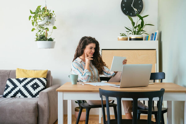 Happy Business Woman Using Digital Tablet at Home. Cheerful smiling businesswoman watching or reading something on her tablet while drinking cup of coffee and sitting at table with documents and laptop computer.