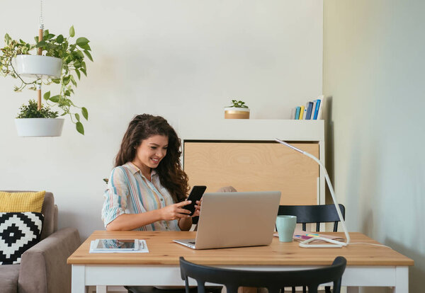 Smiling Businesswoman Using Mobile Phone at Home. Cheerful business woman typing text message on her phone while sitting at desk with a cup of coffee and working on her laptop computer and digital tablet in the living room.