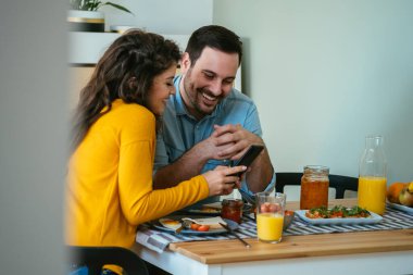 Mutlu çift, evde kahvaltı yaparken cep telefonunu birlikte kullanıyormuş. Masada oturup sağlıklı bir kahvaltı yaparken erkek arkadaşına telefonunu gösteren gülümseyen kız arkadaş..