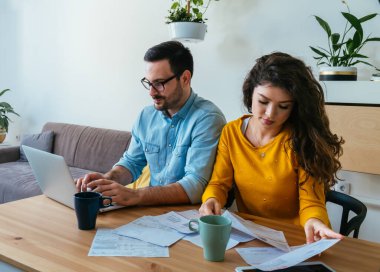 Mutlu Sevgili Çiftler Evlerinde Faturalarını Birlikte Öder. Kız arkadaşı faturaları tutarken ve bir fincan kahve içerken aile bütçesini hesaplarken gülümseyen erkek arkadaş dizüstü bilgisayar kullanıyor..