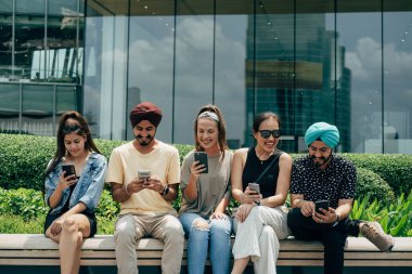 Beş Mutlu Çoklu Etnik Arkadaş Grubu açık havada cep telefonlarını kullanıyor. Gülümseyen arkadaşlar telefonda bir şeyler yazıyor, konuşuyor ve gülüyorlar. Kentteki bankta otururken..