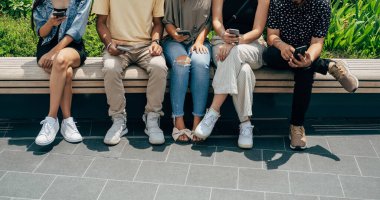 Beş Tanınmayan Çoklu Etnik Arkadaş Grubu cep telefonu kullanıyor ve dışarıda eğleniyor. Arkadaşlarının bir bankta otururken telefonlarına bir şey yazarken ya da izlerken çekilmiş bir fotoğrafı..