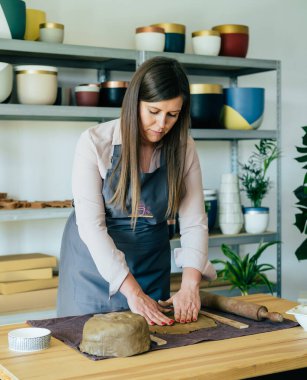 Güzel kadın heykeltıraş atölyesinde Clay 'e baskı yapıyor ve onu şekillendiriyor. Küçük İşletme: Ciddi kadın çömlekçi stüdyosundaki bir masanın üzerindeki kil yığınıyla çalışıyor.