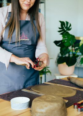 İsimsiz gülümseyen bir kadın atölyesinde Clay 'in Noel süslerini yapıyor. Küçük Sıfır Atık İş: Tanımlanamayan kadın atölyesinde masasında dururken el yapımı Noel süslemeleri için kurabiye keskisi kullanıyor.