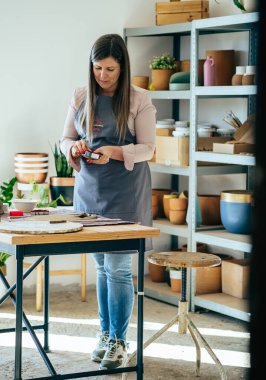 Önlüklü güzel bir kadın Clay 'in Noel süslerini atölyesinde ziyan ediyor. Küçük İş: Ciddi kadın zanaatkar atölyesinde masasında dururken el yapımı Noel süslemeleri için kurabiye keskisi kullanıyor.