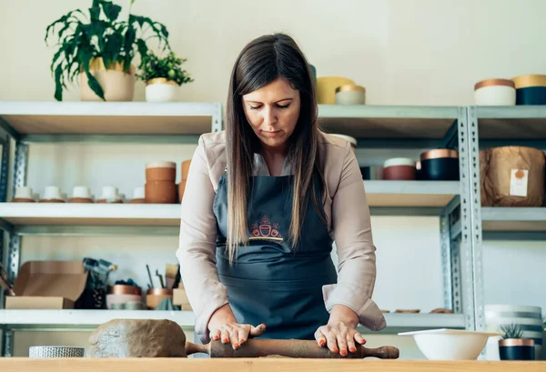 Beautiful Woman Molding Clay At Her Workshop. Small Business: Serious woman potter using rolling pin for clay mass and making ceramics object on a desk in her studio.