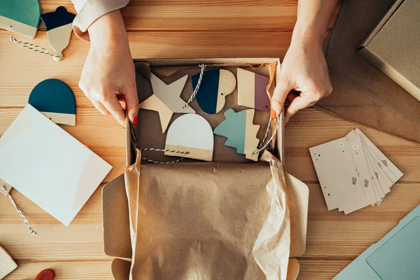 İsimsiz bir kadının dükkanında Clay 'in Noel süslerini paketlediği masa üstü görüntüsü. Küçük Sıfır Atık İş: Atölyesindeki ahşap bir masanın üzerindeki karton bir kutuda teslim edilecek malları paketleyen kadınların el fotoğraflarını kapat.