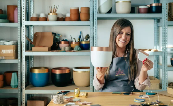 Önlüklü mutlu kadın Potter Clay 'in Noel süslerini atölyesinde ziyan ediyor. Küçük işletme: Gülümseyen kadın, stüdyosunda masasında otururken çiçek saksısı üretiyor ve el yapımı Noel süslemesi yapıyor.