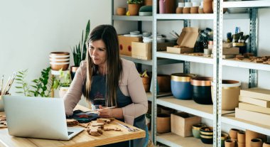 Önlüklü gülümseyen iş kadını dizüstü bilgisayarını kullanıyor ve atölye masasında kilden yapılmış Noel süslerini boyuyor.