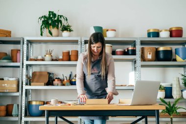 Önlüklü Gülümseyen Kadın 'ın mağazasında Clay' in Noel süslerini teslimata hazırlarken düşük açılı görüntüsü. Mutlu kadın çömlekçi, atölye masasında dizüstü bilgisayarla dururken karton bir kutuda nakliye için mal topluyor..