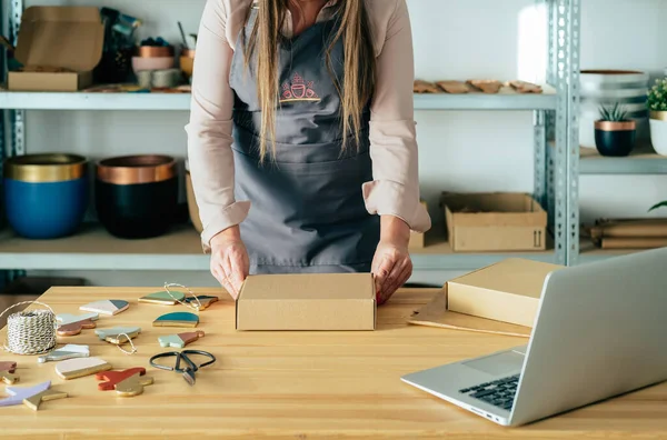 Sustainable Business: Unrecognizable woman potter packing merchandise for shipping in a cardboard box while standing at her workshop desk with a laptop computer