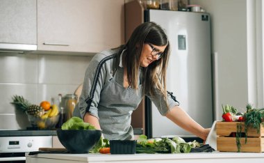 Mutfak masasında yemek pişirirken dijital tablette yemek tarifi okuyan gülümseyen kadın.