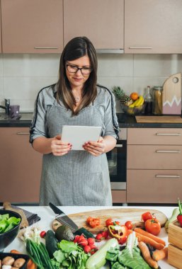 Mutlu ev hanımı, taze organik sebzelerle dolu masasının önünde yemek hazırlarken ve tabletinde sağlıklı yemek dersleri alırken mutlu.
