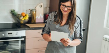 Mutfakta yemek pişirirken dijital tablette yemek tarifi okuyan önlüklü gülümseyen kadın.
