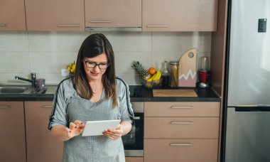 Mutfakta yemek pişirirken dijital tablette yemek tarifi okuyan önlüklü gülümseyen kadın.