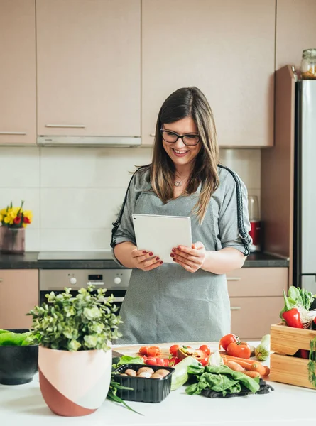 Smiling Woman Reading Recipe On A Digital Tablet While Cooking Lunch In The Modern Kitchen. Happy housewife watching online tutorials for healthy meal on her tablet while standing in front of desk full with fresh organic vegetables and making dinner.