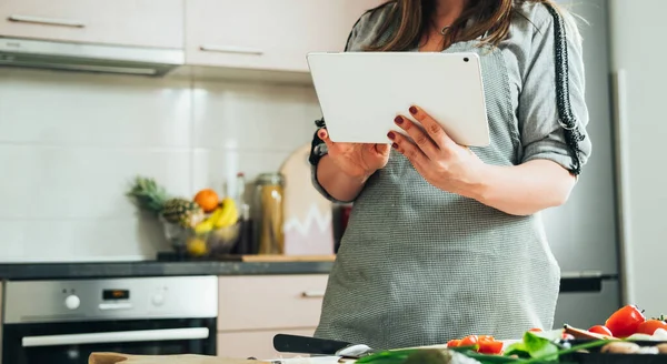 An anonymous woman making healthy lunch with fresh colorful vegetables at the kitchen table while reading recipe online on her tablet