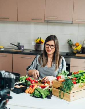 Mutfak masasında oturup yemek kanalı için video çekerken sebze doğrayan gülümseyen kadın.