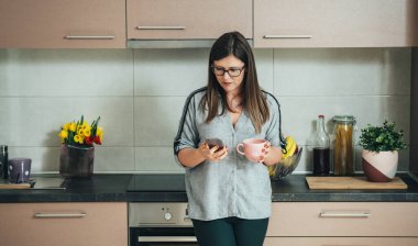Gülümseyen kadın kahve bardağı tutuyor ve mutfakta cep telefonu kullanıyor.