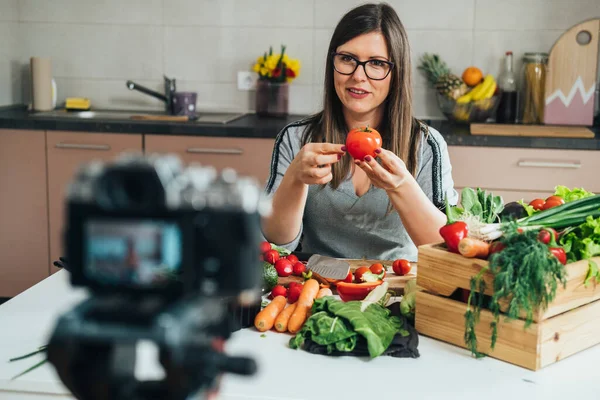 Happy Woman Holding A Tomato While Sitting At Kitchen Desk And Filming Video For Her Food Channel