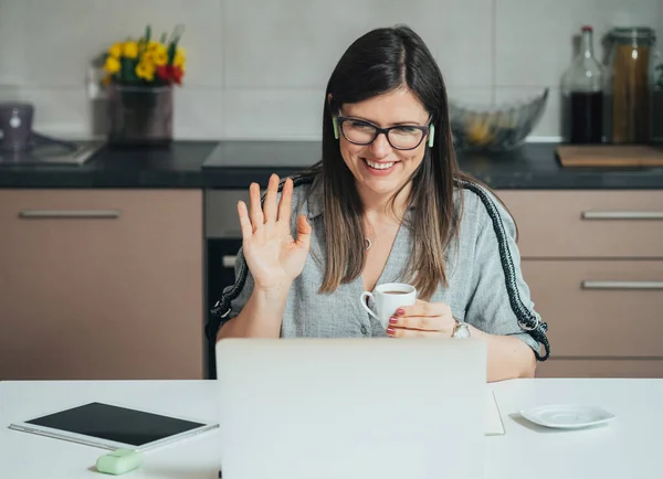 Smiling Businesswoman Waving And Say Hello To Colleagues At Web Conference Meeting On A Laptop Computer While Drinking Cup Of Coffee In The Kitchen