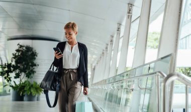 Low Angle View Of Happy Business Woman Holding Shopping Bags And Using Mobile Phone While Walking On A Footbridge