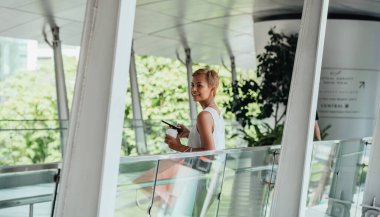 Happy Woman Using Mobile Phone And Drinking Cup Of Coffee While Holding Shopping Bags And Standing At Modern Balcony