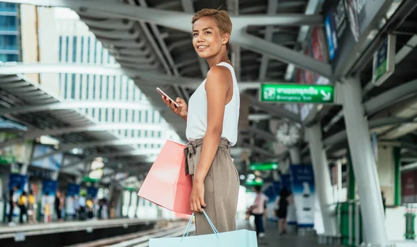 Happy Woman Holding Shopping Bags And Using Mobile Phone While Standing At Railway Platform