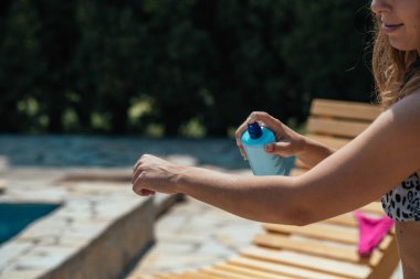 Unrecognizable young woman spraying suntan lotion on her arm at the swimming pool