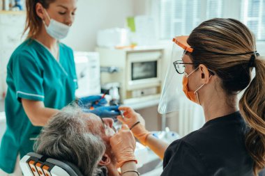 Diş hekiminde dişçi asistanı dişçiye dişçi aletini verirken, dişçi koltuğunda oturan tanınmayan bir erkek hasta diş hekimine...