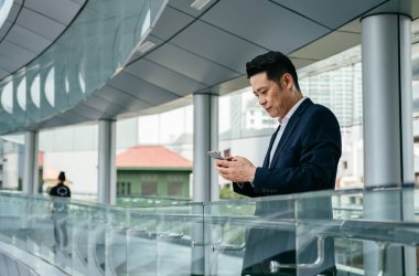 Modern bir balkonda duran ve cep telefonuna mesaj yazan zarif mavi takım elbiseli gülümseyen Asyalı iş adamı.