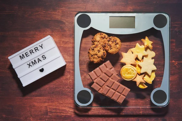  Işık kutusunda mutlu noeller yazıyor. Çikolata, kurabiye ve noel tatlısıyla kilo ölçeği. İnsanlar Noel 'de kilo alırlar..                            