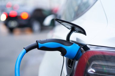  Beyaz Elektrikli araba şehirdeki otoparkta şarj oluyor. Gri bir günde şehir manzarası. Arka planda bulanık araba.                            