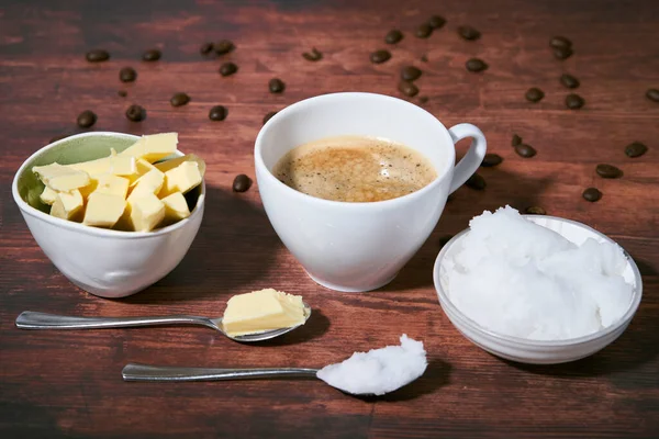 Bulletproof coffee in a white cup. Coconut oil, butter and coffee beans can be seen. Spoons in front of the cup. Close up. Bulletproof coffee is part of the ketogenic diet.                              