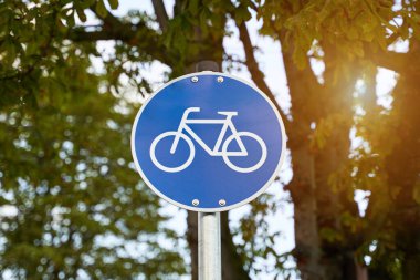 Bisikletçiler için yol tabelası. Mavi tabelada motosiklet görünüyor. Arka planda görünen odak dışı doğa. Çevresel bilinç hareketi. Güneş ağaçların arasından parlıyor.                               