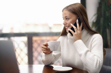 Genç bir kadın kahve içiyor ve telefonla konuşuyor.