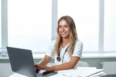 Bilgisayarlı gülümseyen bir kadın doktorun portresi tıp ofisinde oturuyor. Sağlık hizmetleri kavramı