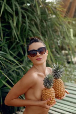 Tropikal arka planda poz veren genç ve güzel bir kız, elinde ananaslarla. Bikinili bronz tenli kadın.. 